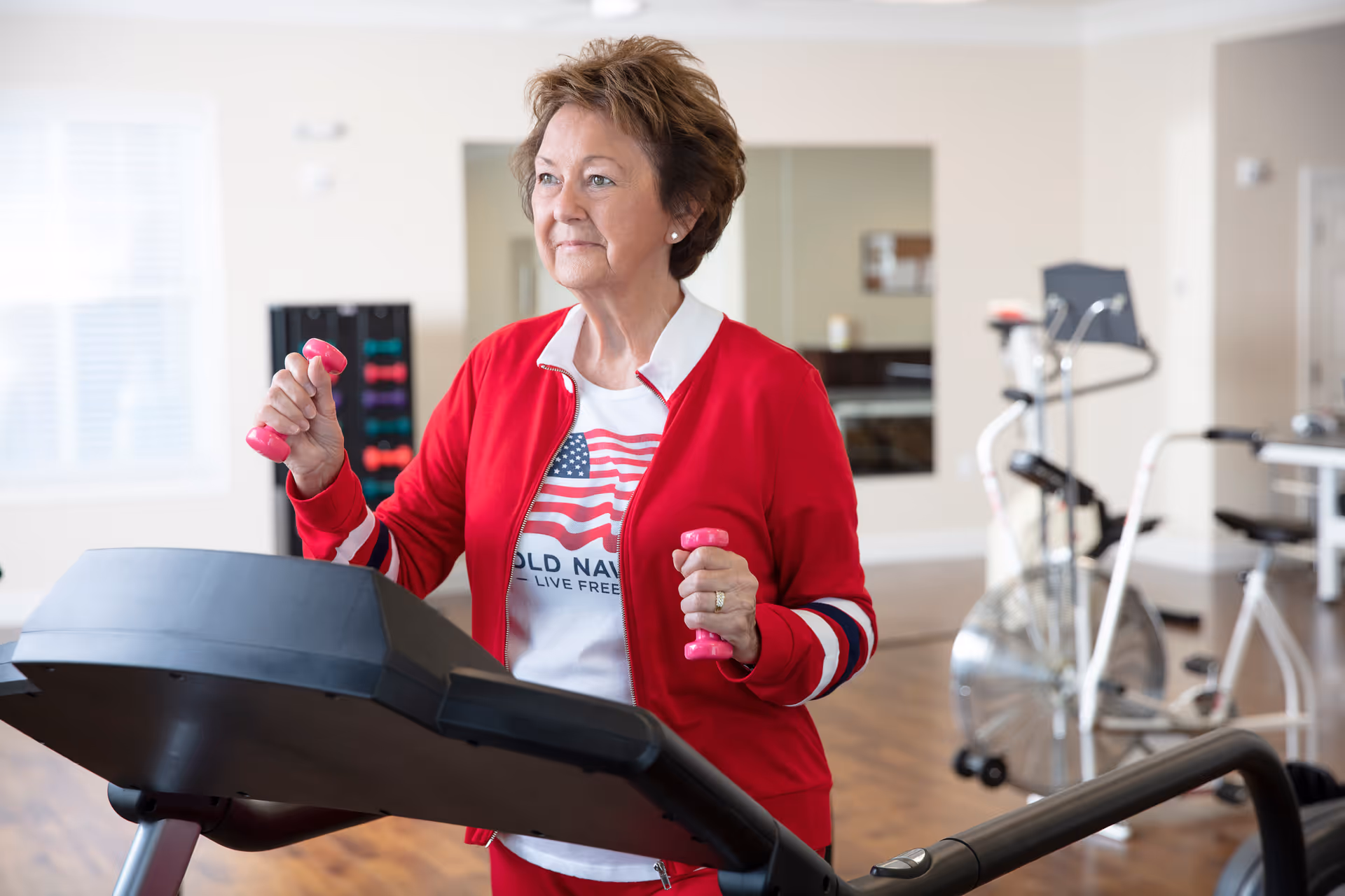 An elderly woman wearing a red jacket and white t-shirt with an American flag design is exercising on a treadmill while holding small pink dumbbells in a well-lit fitness room with exercise equipment in the background.