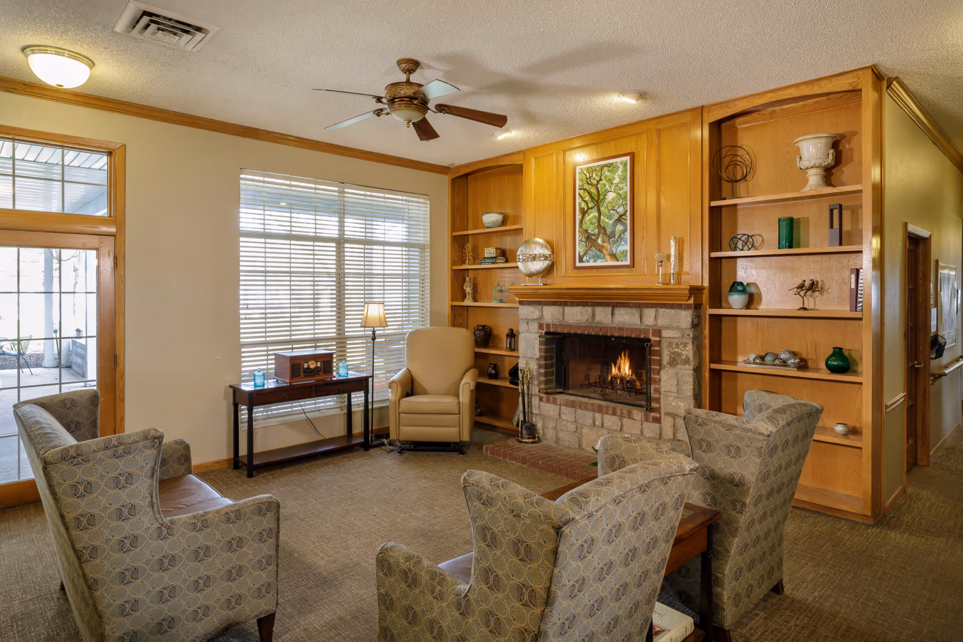 A cozy living room with a stone fireplace surrounded by wooden built-in shelves displaying decorative items. The room has four patterned armchairs arranged around a small wooden table, a beige armchair near a window with blinds, a floor lamp, and a small table with a vintage radio. A ceiling fan is mounted on the white ceiling, and a glass door leads to an outdoor area.