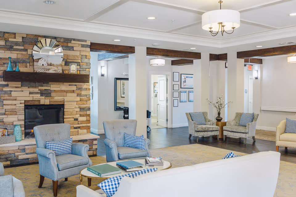 Bright assisted living facility lounge with armchairs and sofas arranged around a stone fireplace and coffee table.
