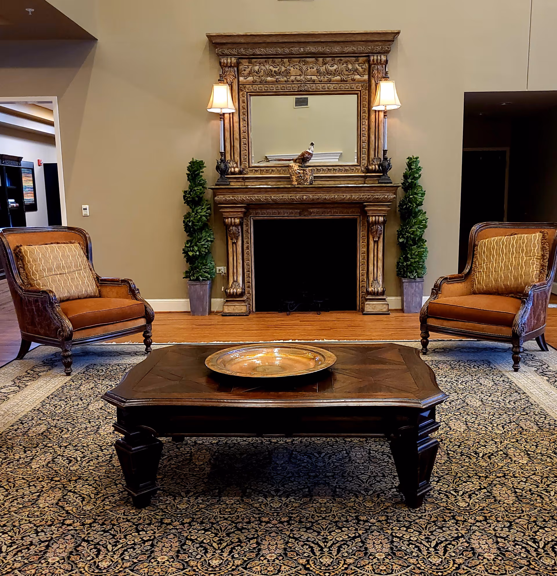 A furnished lounge with a decorative fireplace and mirror, two upholstered armchairs, and a wooden coffee table on a patterned rug.