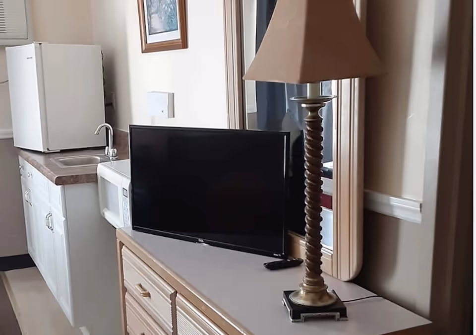 Small studio-style room with a kitchenette (sink, microwave, mini-fridge), a TV on a dresser, and a tall table lamp in front of a mirror.