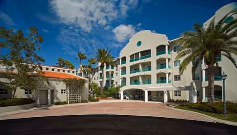 White, multi-story senior living building with balconies, palm trees, and a circular driveway.