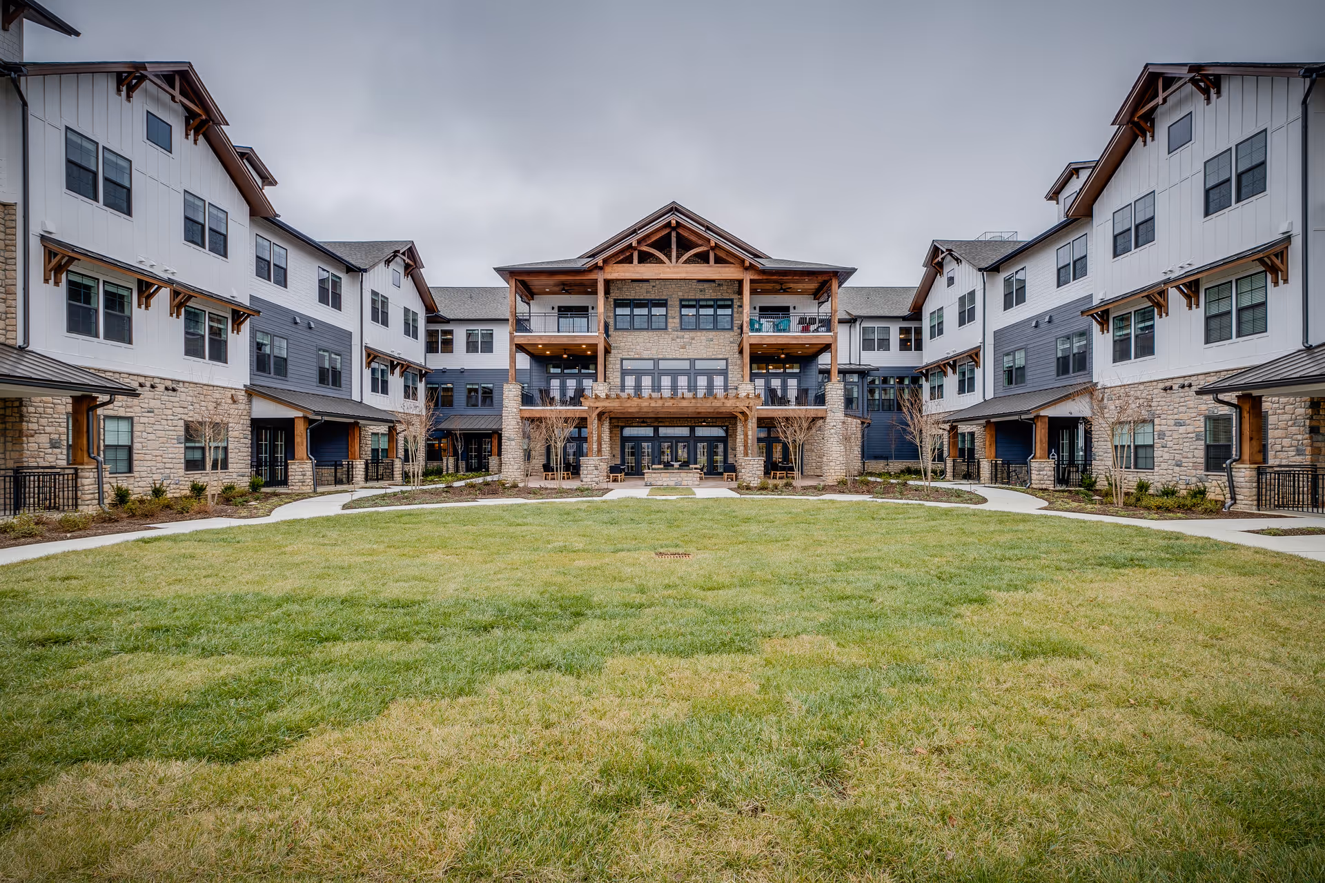 Exterior view of a large, modern senior living facility with three stories, featuring a combination of stone and siding on the facade. The building surrounds a spacious, well-maintained grassy courtyard with paved walkways and small trees. The sky is overcast.