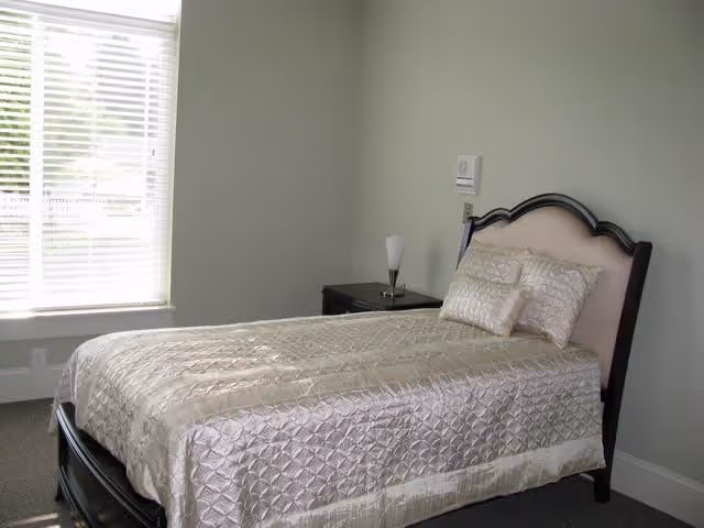A neatly made single bed with a light-colored quilted bedspread and matching pillows in a simple bedroom with a window covered by white blinds and a small bedside table with a lamp.