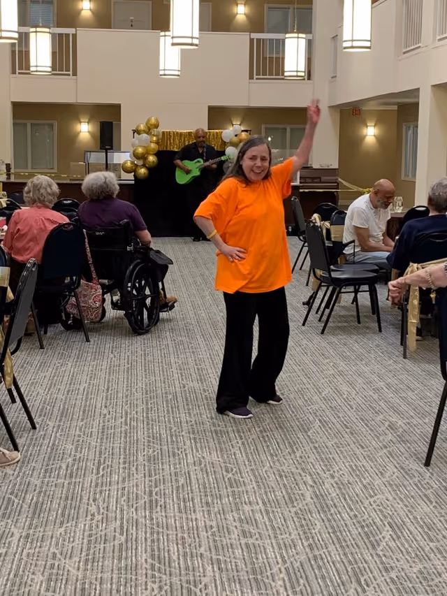 A woman in an orange shirt is dancing and smiling in a large room with carpeted floor. Behind her, a man is playing a green guitar on a small stage decorated with gold and white balloons. Several elderly people are seated at tables and in chairs around the room, some watching the performance.