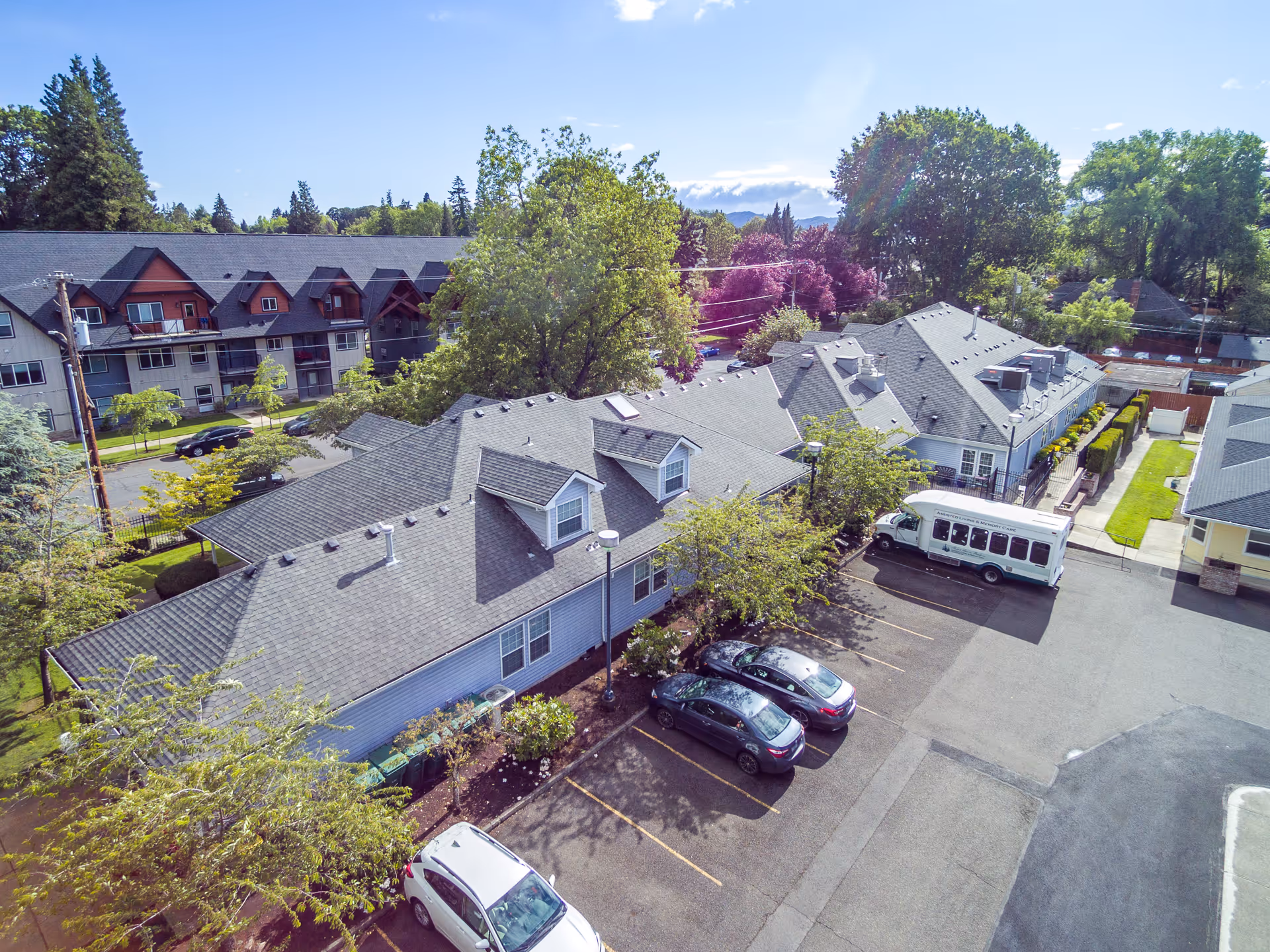 Aerial view of a memory care facility's buildings with a parking lot, several parked cars and a white shuttle bus, surrounded by trees and neighboring residences.