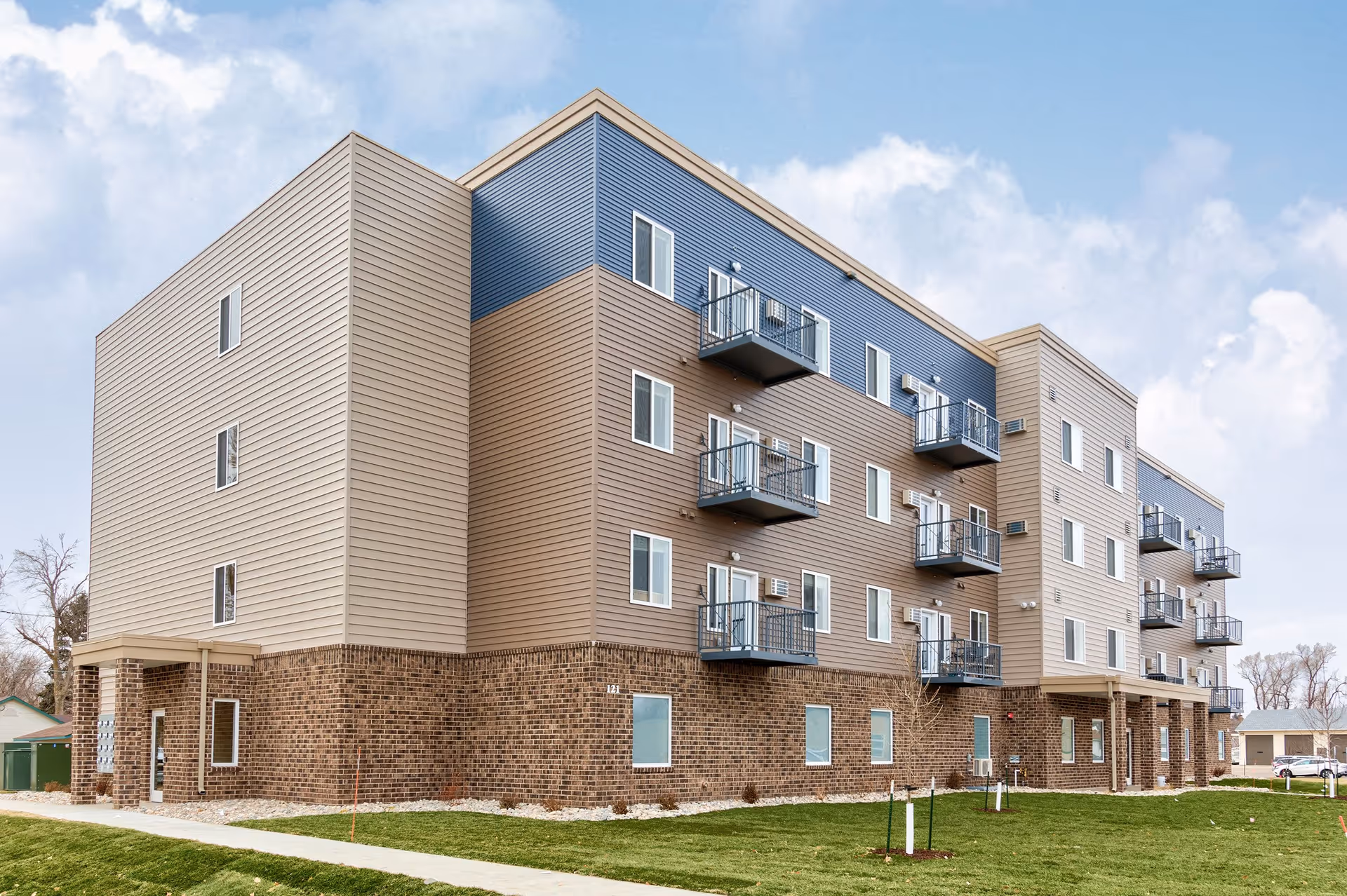 Exterior view of a modern multi-story residential building with a combination of brick and siding facade, featuring small balconies on the upper floors and a well-maintained lawn in the foreground under a partly cloudy sky.