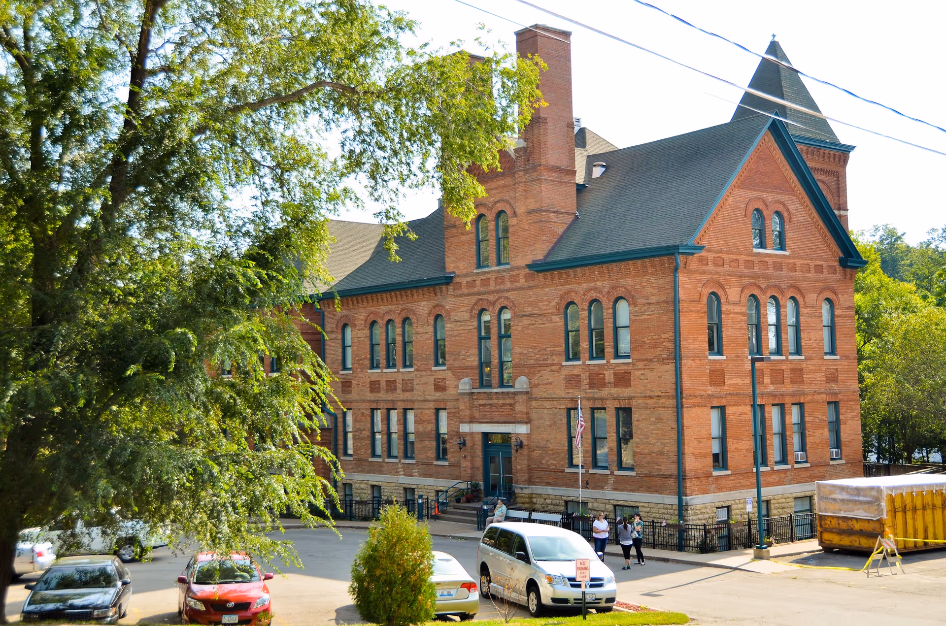 A large, historic brick building with multiple windows and a steep roof, surrounded by trees and parked cars. There are a few people standing near the entrance, and an American flag is visible near the door.
