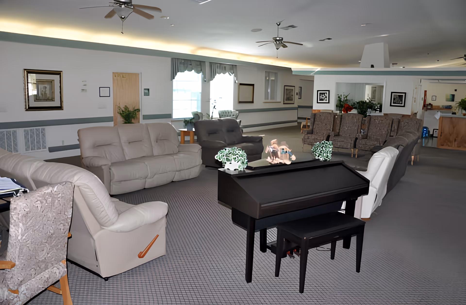 Spacious assisted-living common lounge with sofas, armchairs, a piano, and decorative plants.