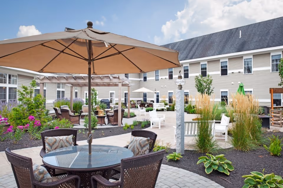 Outdoor courtyard area at Sunrise of Huntington Common featuring a round glass table with four wicker chairs and a large beige umbrella. The courtyard is landscaped with various plants, flowers, and shrubs, and includes additional seating areas with white benches and wicker chairs. The building surrounding the courtyard has beige siding and multiple windows under a dark shingled roof, with a partly cloudy blue sky overhead.