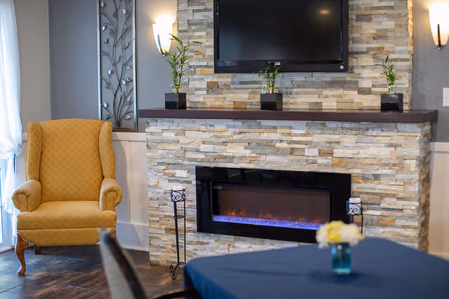 A cozy living room area featuring a yellow upholstered armchair to the left, a stone wall with a modern electric fireplace below a mounted flat-screen TV, and a dark wooden mantel with three small potted plants. In the foreground, there is a table covered with a dark blue tablecloth and a small vase with yellow and white flowers.