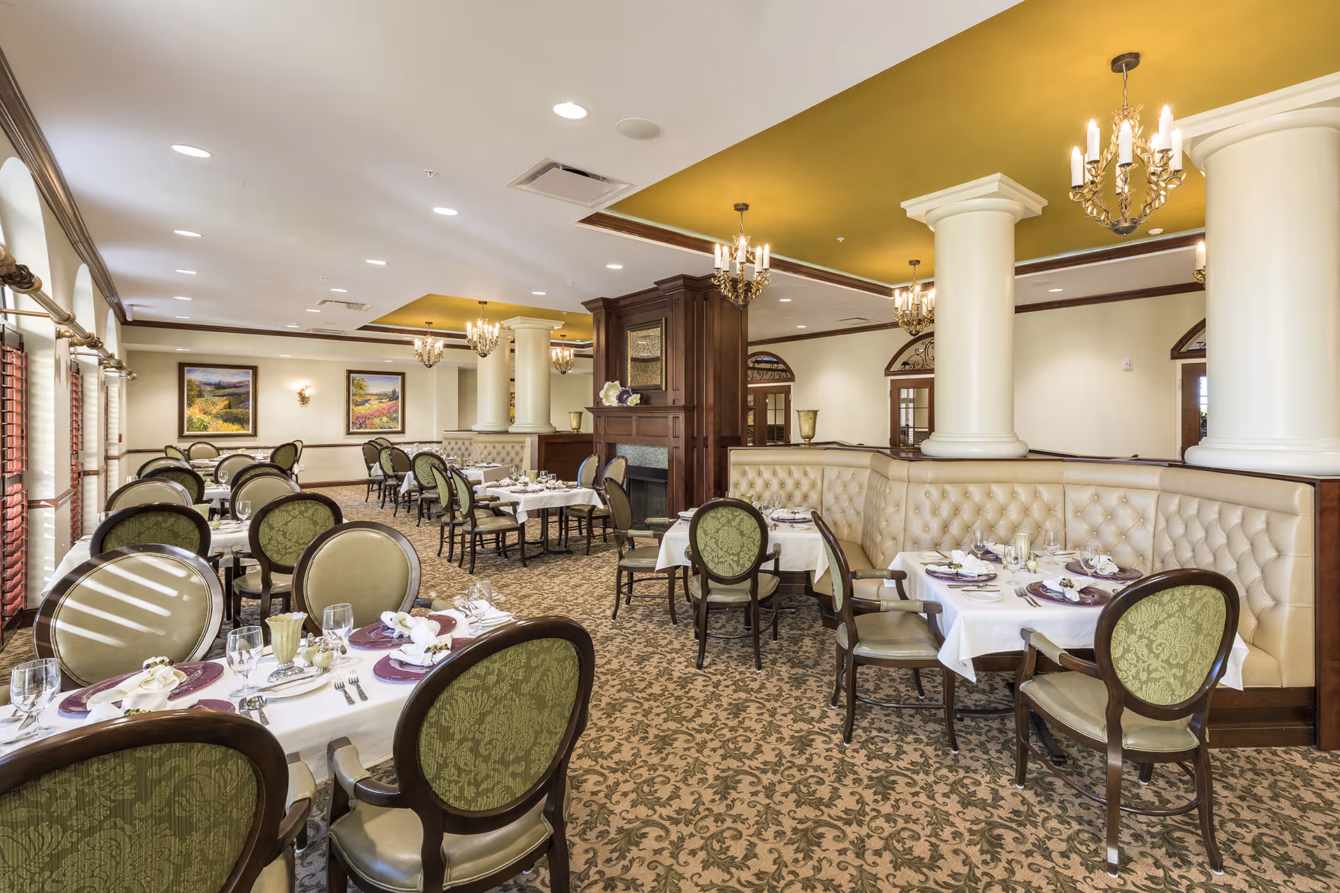 Elegant dining room with round tables set for service, green upholstered chairs, tufted banquettes, columns and chandeliers.