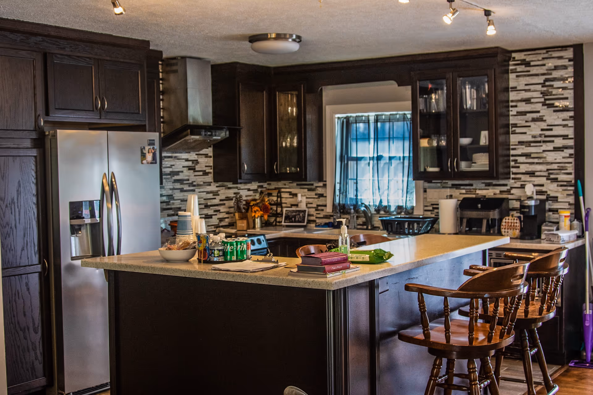A modern kitchen with dark wooden cabinets and a light-colored countertop island. The kitchen features a stainless steel refrigerator, a stove with a range hood, a window with blue curtains, and various items on the counter including books, soda cans, a bowl, and hand sanitizer. There are two wooden bar stools at the island.