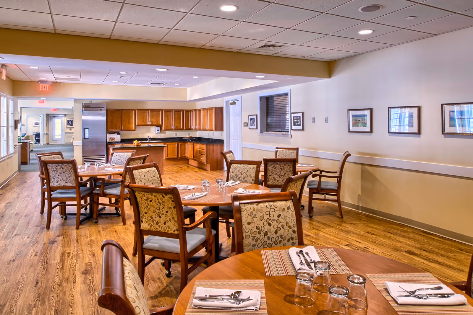 Bright dining room with round wooden tables and upholstered chairs set for meals and an open kitchen area in the background.