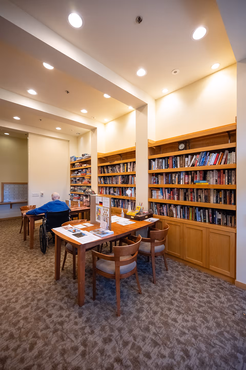 A bright communal library area with built-in bookshelves, a wooden table with chairs, and a resident in a wheelchair seated at the table.
