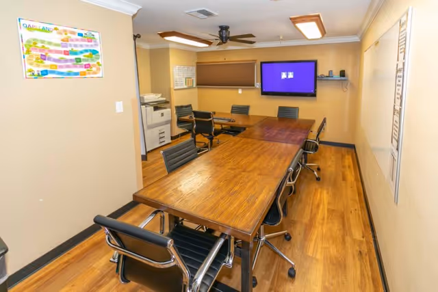 A long interior conference room with a wooden table, rolling chairs, wall-mounted TV, and office equipment.