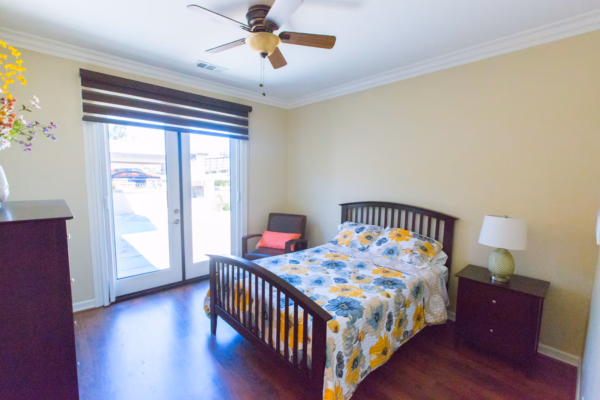 A bright bedroom with a wooden bed frame and a floral bedspread in yellow, blue, and gray tones. There is a wooden nightstand with a lamp on the right side of the bed and a wooden dresser on the left side of the room. A comfortable armchair with a coral cushion is placed in the corner near glass sliding doors that lead outside. The room has light-colored walls, a ceiling fan with wooden blades, and dark wood flooring.