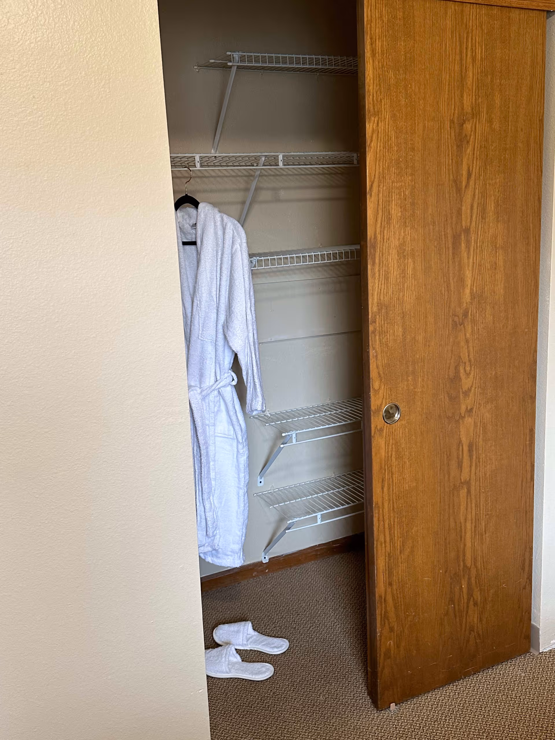 Partially open wooden closet door revealing wire shelving inside. A white bathrobe is hanging on a black hanger on the left side of the closet, and a pair of white slippers are placed on the carpeted floor in front of the closet.