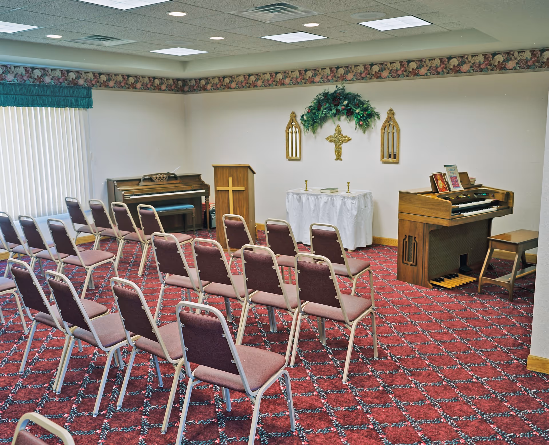 Small chapel-style room with rows of chairs on a red patterned carpet facing a lectern, altar table, piano and organ.