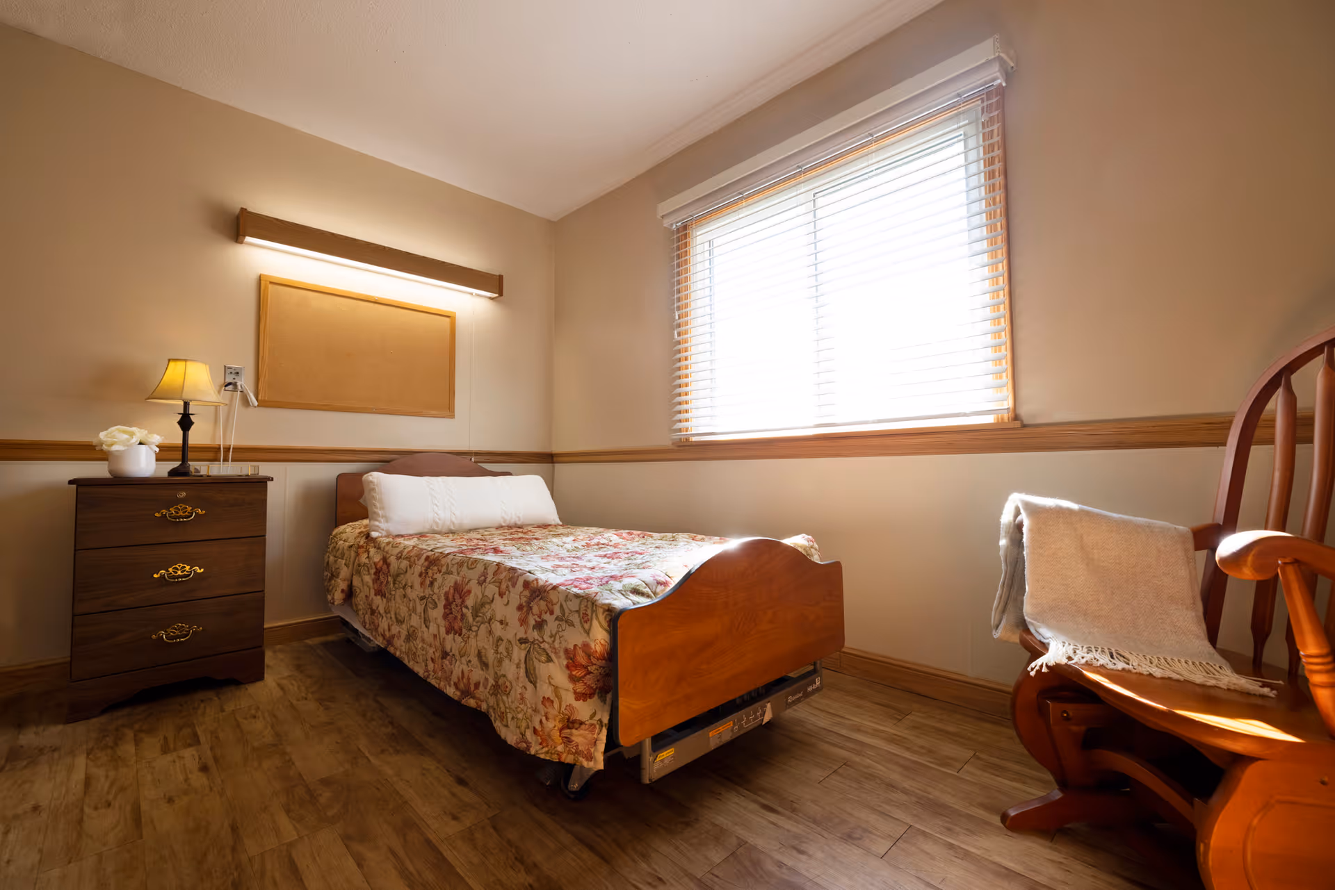 Sunlit small bedroom with a single wooden bed covered by a floral quilt, a nightstand with a lamp and flowers, and a wooden rocking chair by a window with blinds.