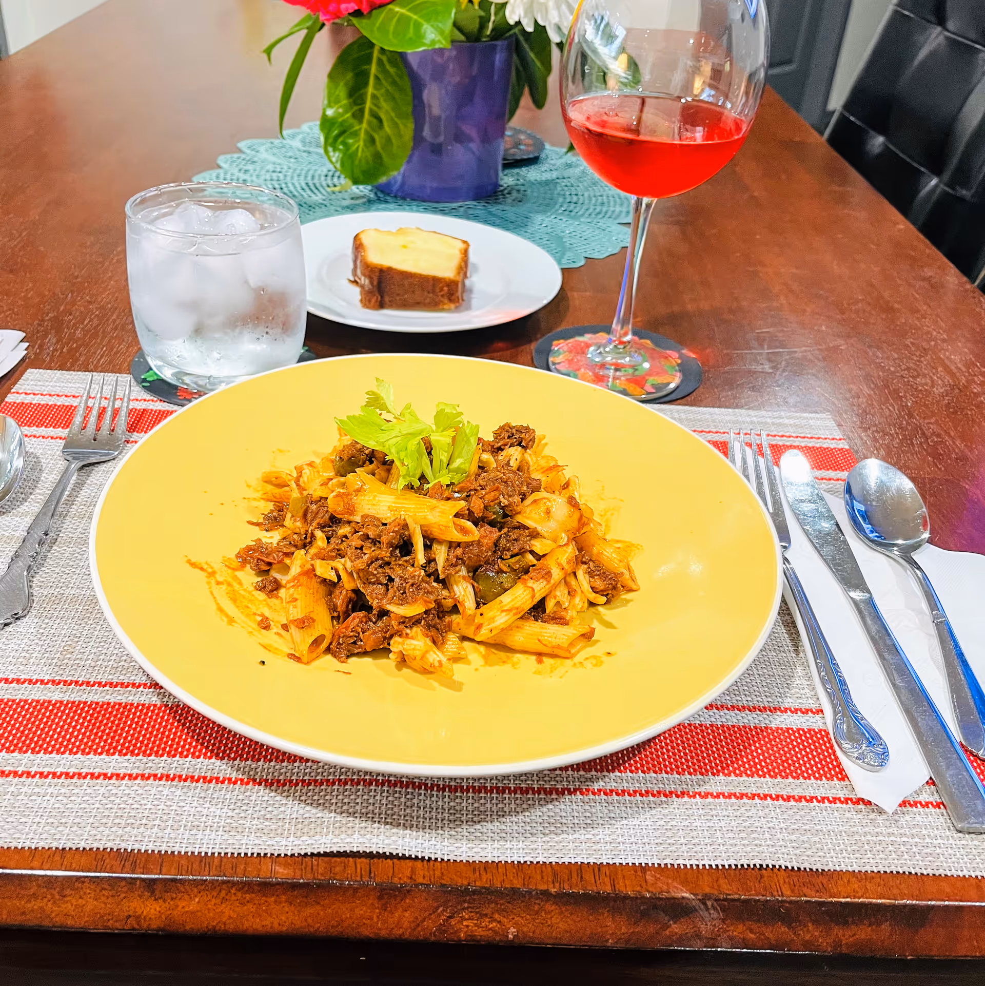 A dining table set with a yellow plate of pasta topped with celery, a glass of ice water, a slice of cake on a white plate, and a glass of red beverage. There is a purple flower pot with green leaves and pink flowers in the background on a blue doily.