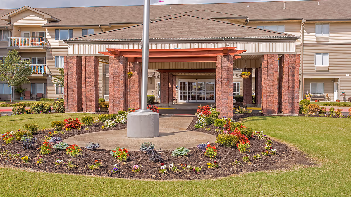 Front entrance of a multi-story senior living building with a covered porte-cochere, flagpole, and landscaped flower beds.