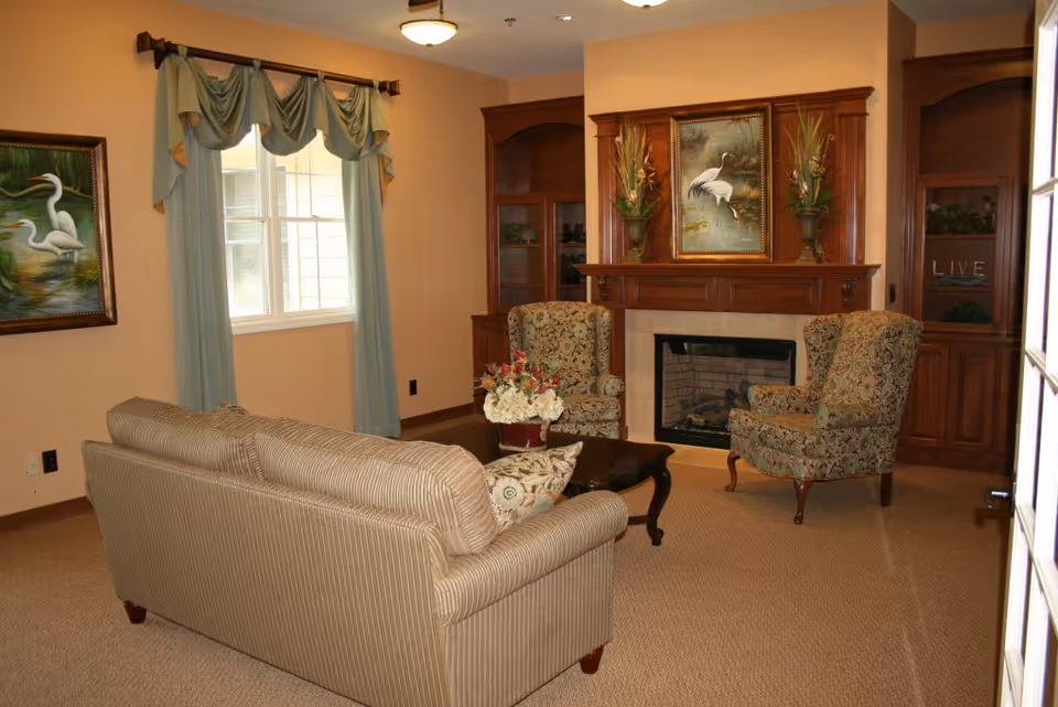 A cozy living room with a sofa, two patterned armchairs, a fireplace with a wooden mantel and artwork, and a window with drapes.