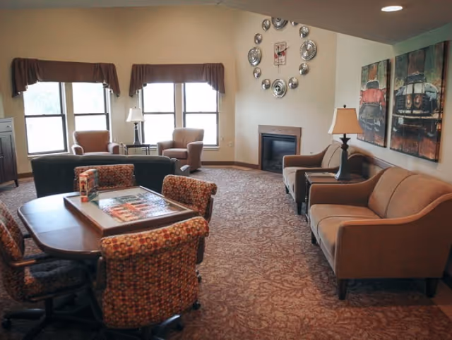 Communal senior living lounge with sofas, armchairs, a game table with chairs, a fireplace, and wall art.