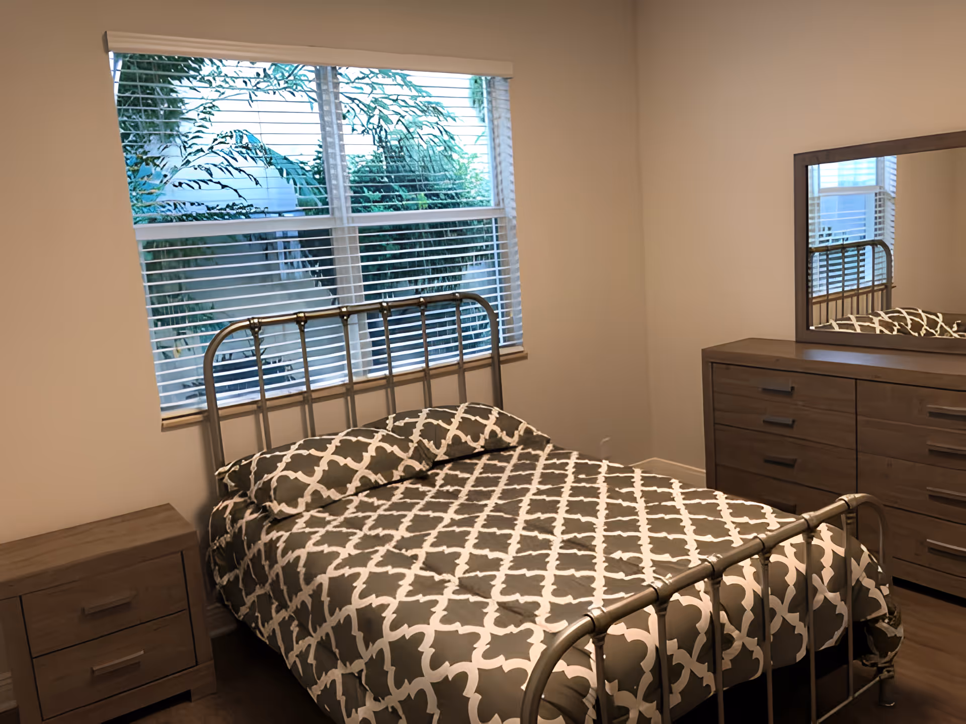 A bedroom with a metal bed frame and a bed covered with a gray and white patterned comforter and matching pillows. There is a wooden nightstand with two drawers to the left of the bed and a wooden dresser with six drawers and a large mirror on the right side of the room. A window with white blinds shows greenery outside.