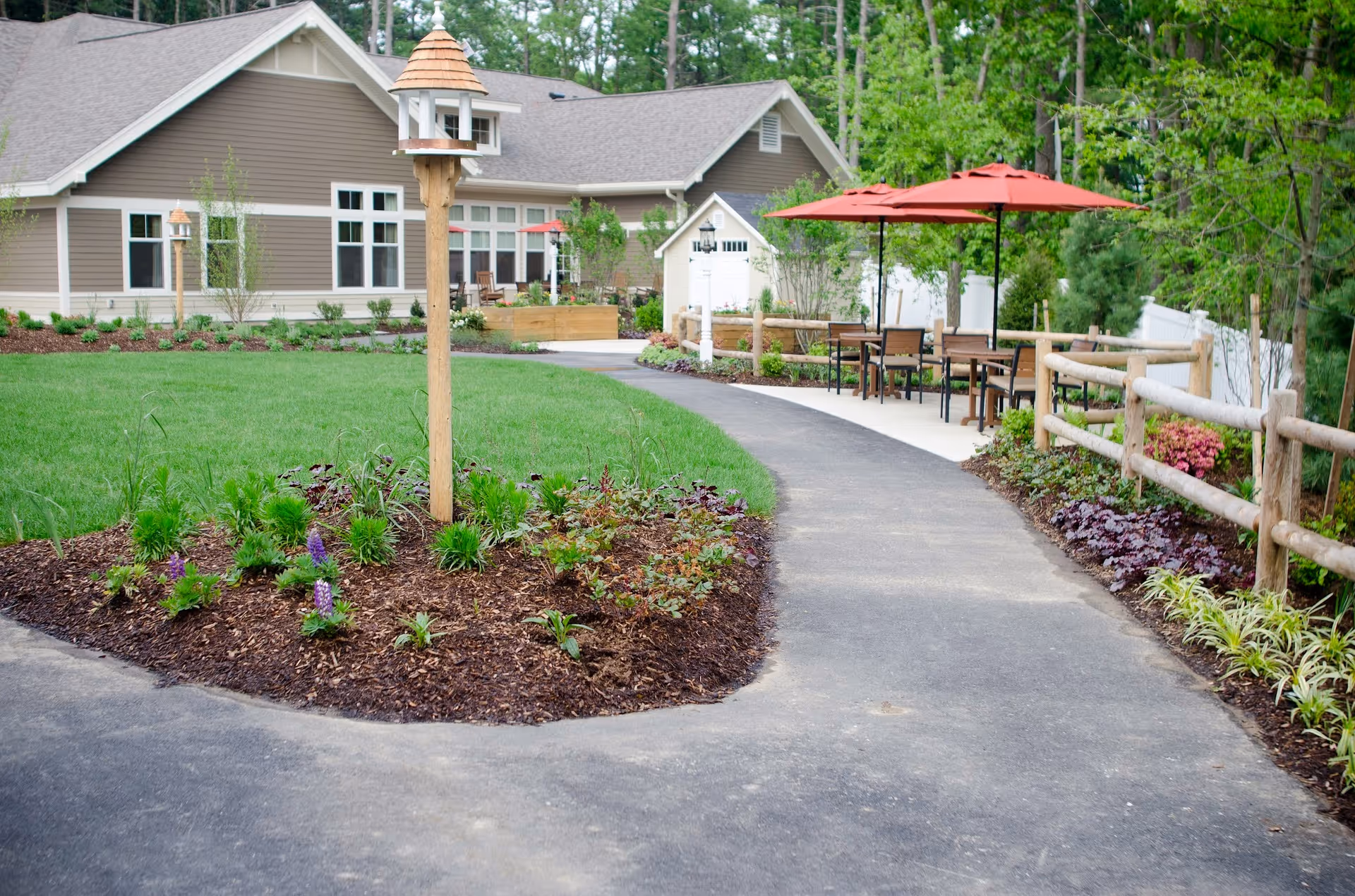 Landscaped outdoor courtyard with a paved walkway, a wooden birdhouse, patio tables under red umbrellas, and a building in the background.