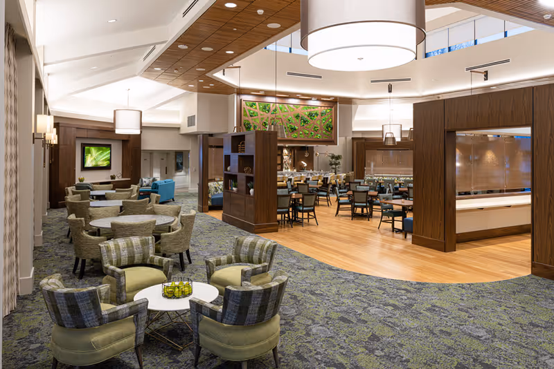 A spacious and well-lit common area in a senior living facility featuring multiple seating arrangements including round tables with chairs and cushioned armchairs around small white tables. The area has a mix of carpet and wooden flooring, modern pendant lighting, and decorative wooden panels. In the background, there is a dining area with tables and chairs, and a green wall art piece above a wooden divider.
