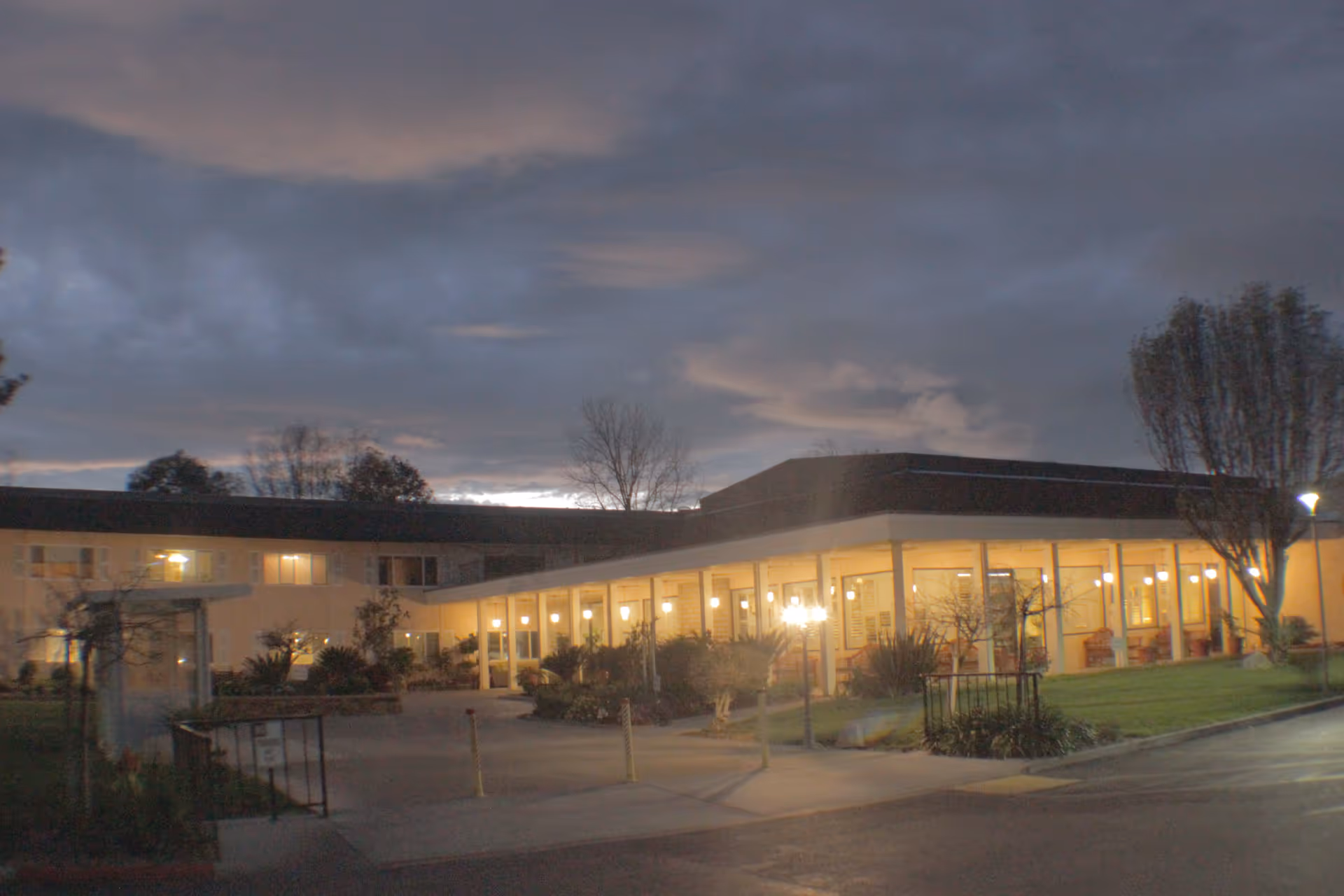 Exterior view of The Californian senior living facility at dusk with lights glowing from inside the building, a covered walkway, trees, and a cloudy sky.