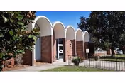 Front entrance of a brick building with white arched canopy elements, columns, and surrounding trees and lawn.