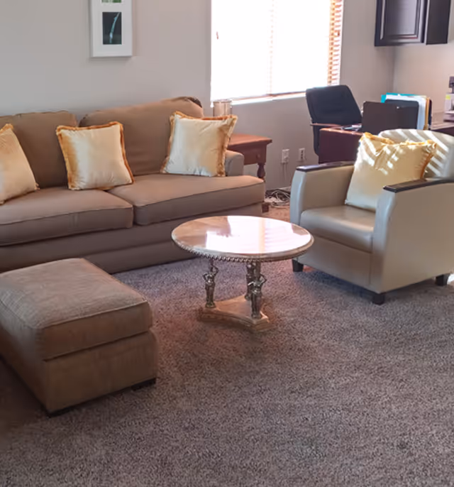 A cozy living room with a beige sofa adorned with four cream-colored pillows, a matching beige armchair with a similar pillow, a cushioned ottoman, and a round marble-top coffee table on a carpeted floor. In the background, there is a window with blinds, a wooden side table, and an office chair with a desk and some office supplies.