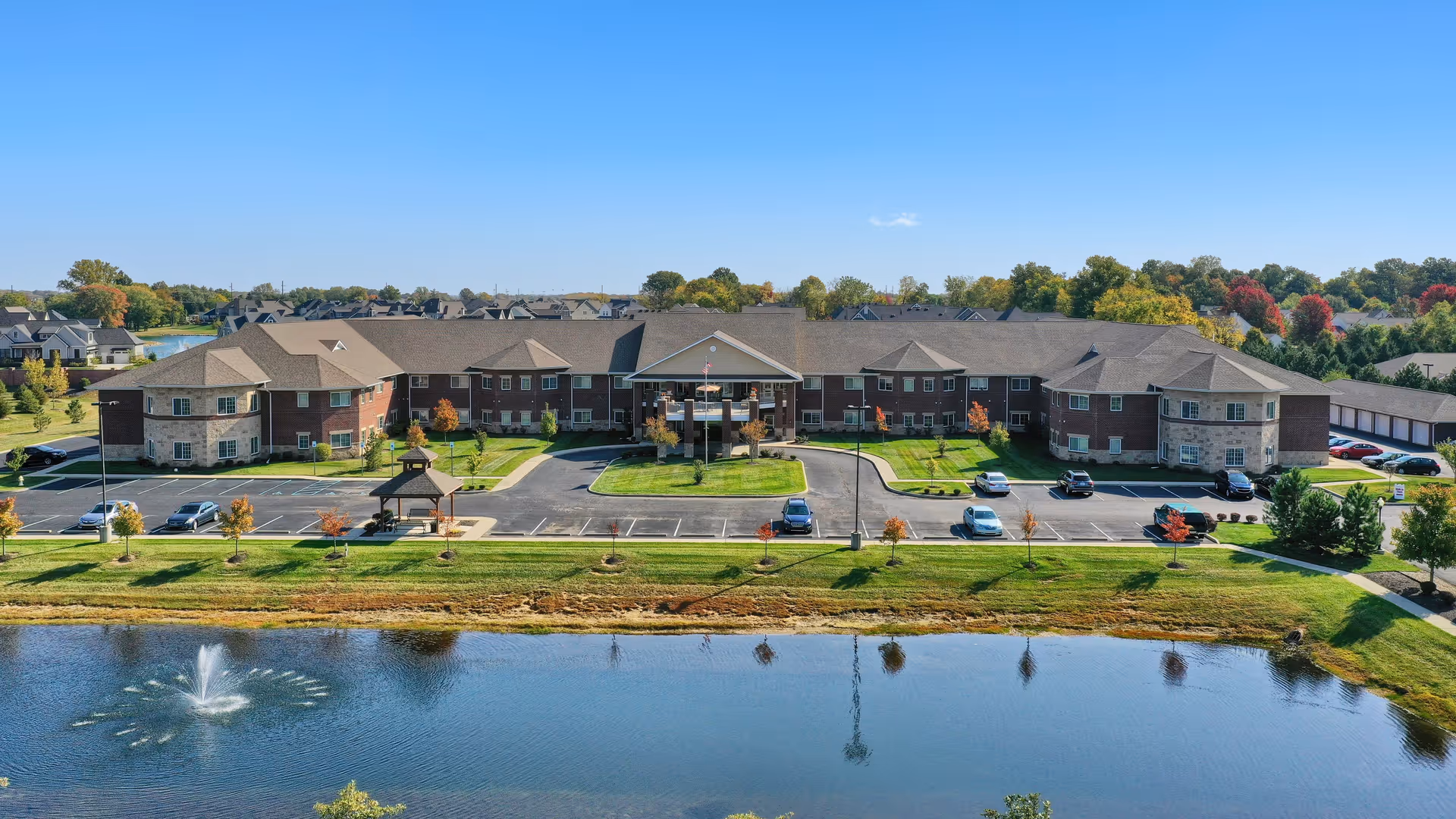Aerial view of Magnolia Springs Bridgewater senior living facility showing a large two-story building with a peaked entrance, surrounded by a parking lot with several cars. In the foreground, there is a pond with a water fountain, and the background features trees with autumn foliage under a clear blue sky.