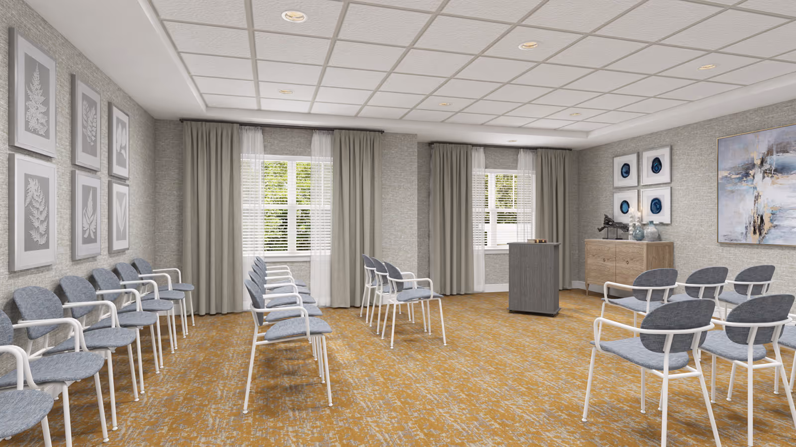 A spacious interior meeting room with rows of gray chairs arranged facing a small podium, neutral walls, artwork, and windows with curtains.