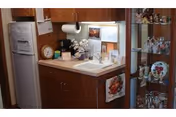 A small kitchenette area with a white refrigerator, wooden cabinets, a sink, and a countertop. There are various items on the counter including a clock, a coffee maker, and decorative flowers. To the right, there is a glass display cabinet filled with decorative glassware and figurines.