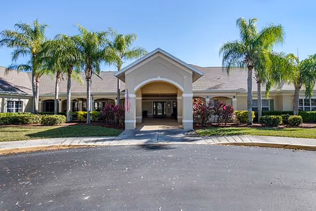 Front exterior view of a single-story building with a covered entrance, surrounded by palm trees and landscaped bushes under a clear blue sky.