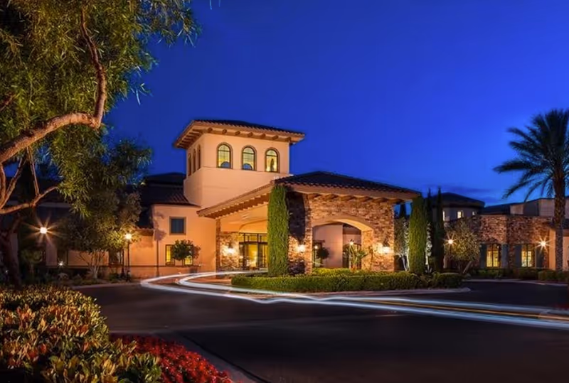Exterior view of a senior living facility building at dusk with warm lights illuminating the entrance. The building features a Mediterranean architectural style with stone and stucco walls, arched entryways, and a tower with arched windows. Surrounding the building are trees, shrubs, and palm trees under a deep blue evening sky.