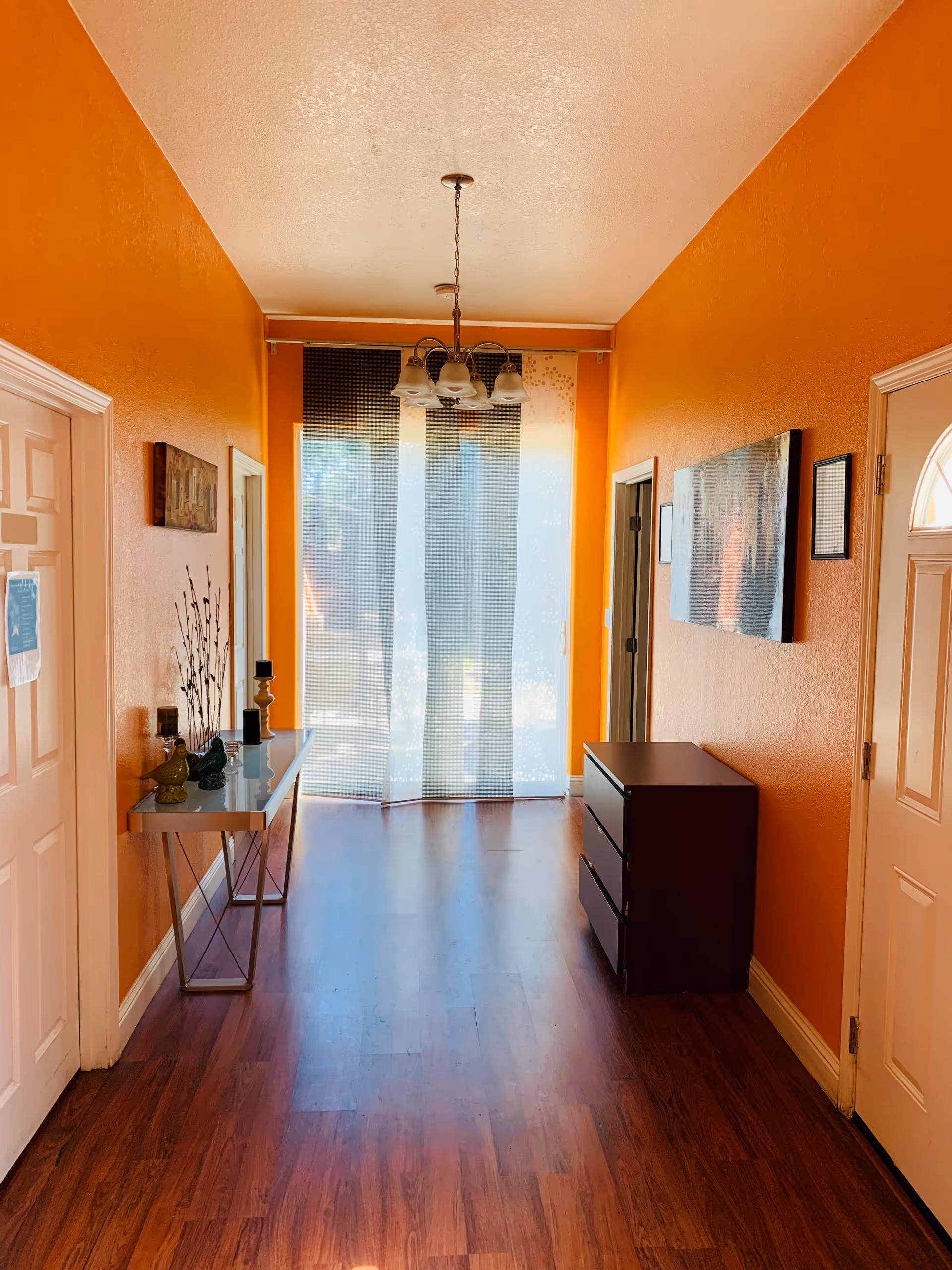 A hallway with bright orange walls and wooden flooring. On the left side, there is a white door with a paper attached to it and a narrow glass table with decorative items including candles and vases. On the right side, there is a dark wooden chest of drawers and a wall-mounted artwork. At the end of the hallway, there is a large window covered with sheer curtains, allowing natural light to fill the space. A ceiling light fixture with multiple lamps hangs in the center of the hallway.