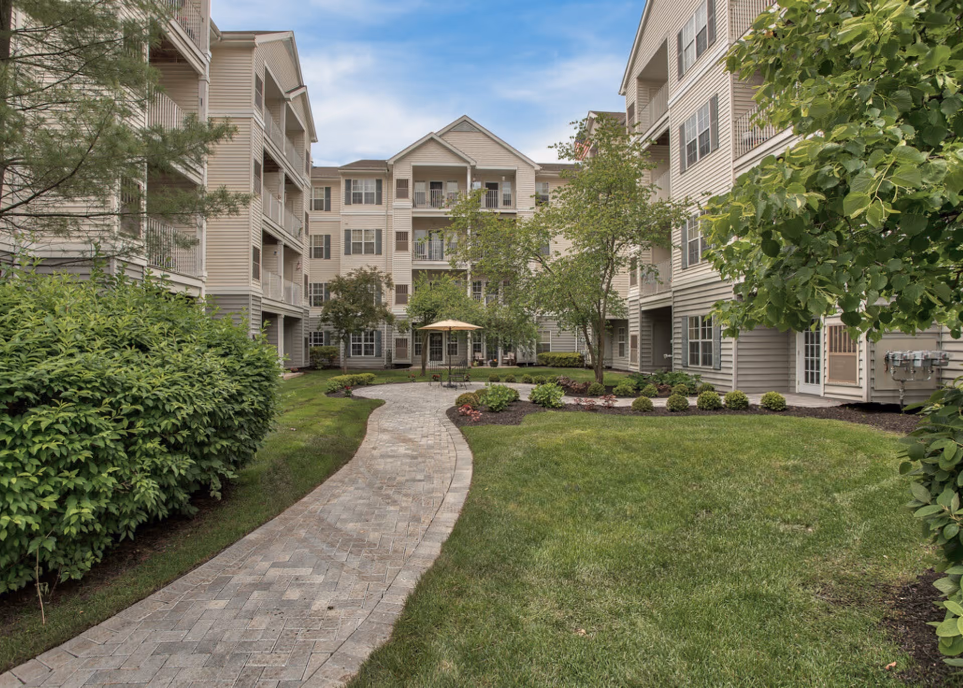Brick paver walkway through a landscaped courtyard between multi-story beige apartment buildings with balconies.