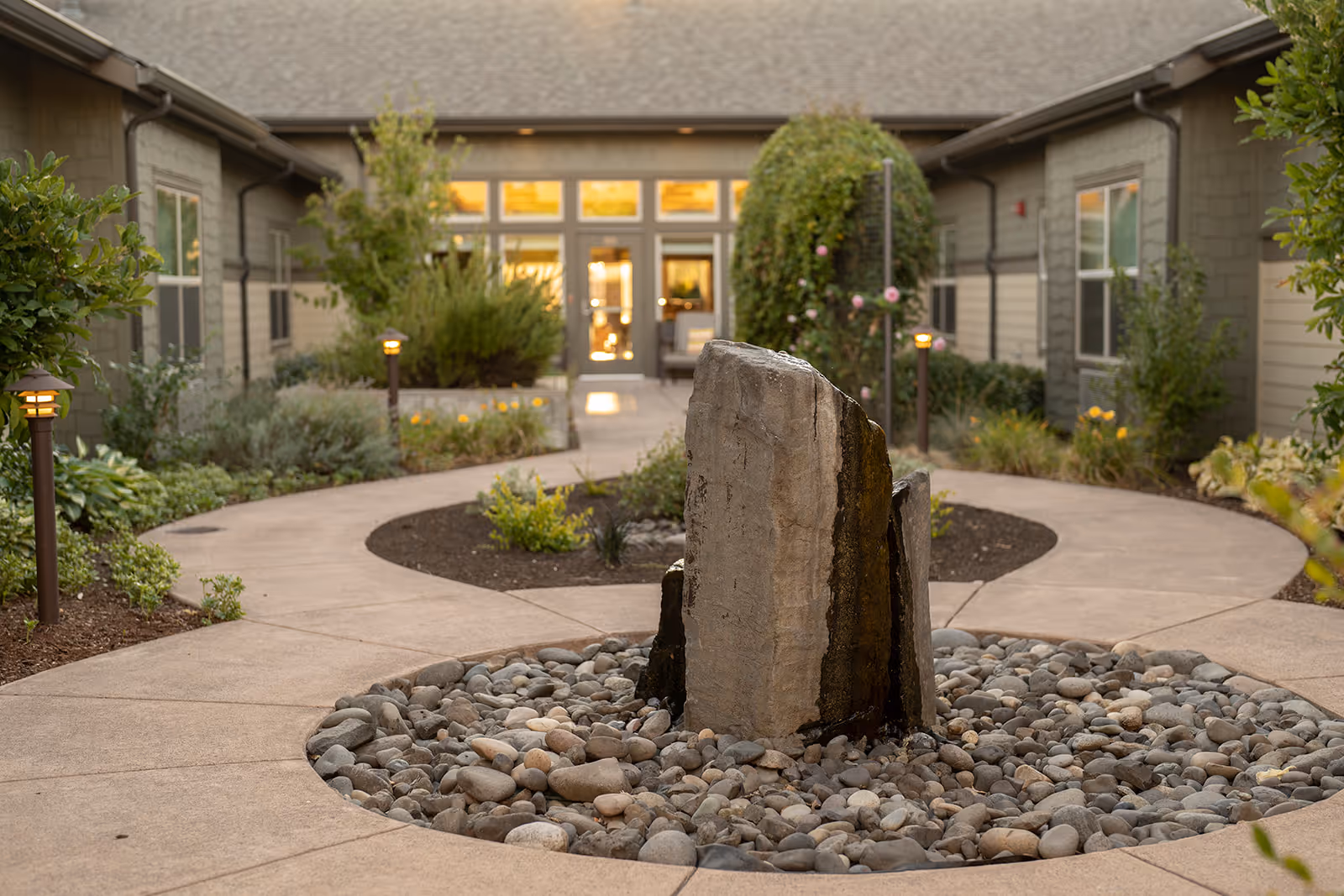 A landscaped outdoor courtyard area with a circular stone water feature in the center surrounded by a paved walkway. The courtyard is bordered by plants and shrubs, with a building featuring multiple windows and a door in the background.