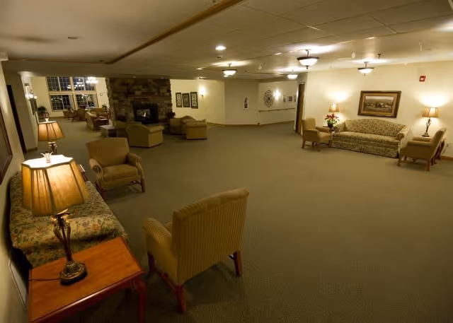 A spacious living room area in an assisted living facility with multiple armchairs and sofas arranged around a stone fireplace. The room is softly lit with table lamps and ceiling lights, and decorated with framed artwork and a flower arrangement on a side table.