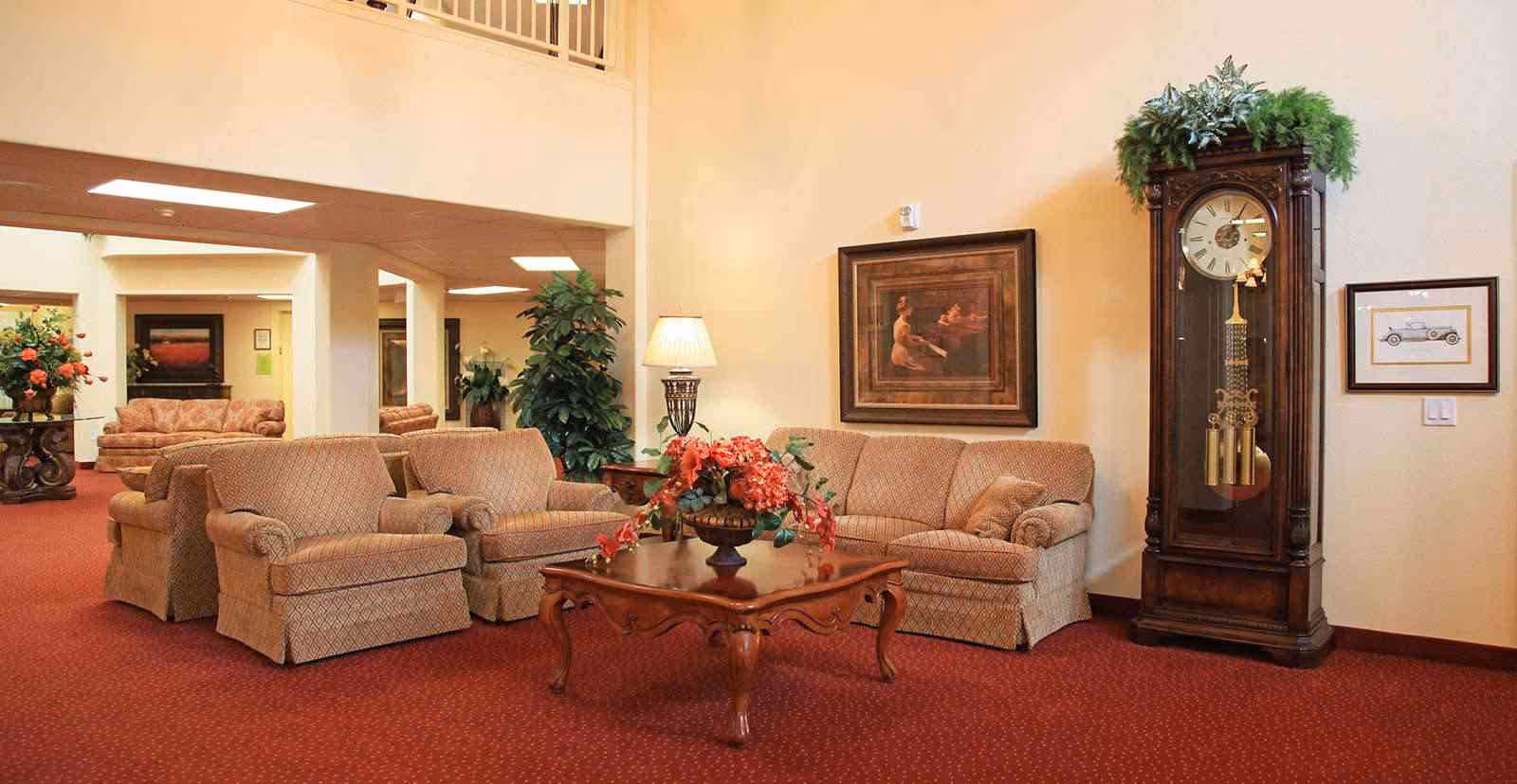 A cozy living room area with beige upholstered armchairs and a sofa arranged around a wooden coffee table with a floral centerpiece. The room has red carpet flooring, a tall grandfather clock with plants on top, framed artwork on the walls, and a table lamp providing warm lighting.
