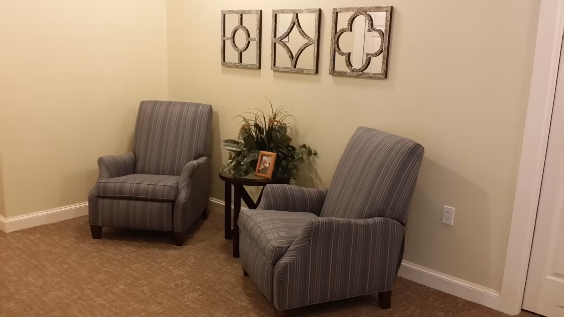 A cozy corner with two gray striped armchairs separated by a small round wooden table holding a green plant and a framed photo. Three decorative wooden wall hangings with geometric patterns are mounted above the chairs on a light-colored wall.