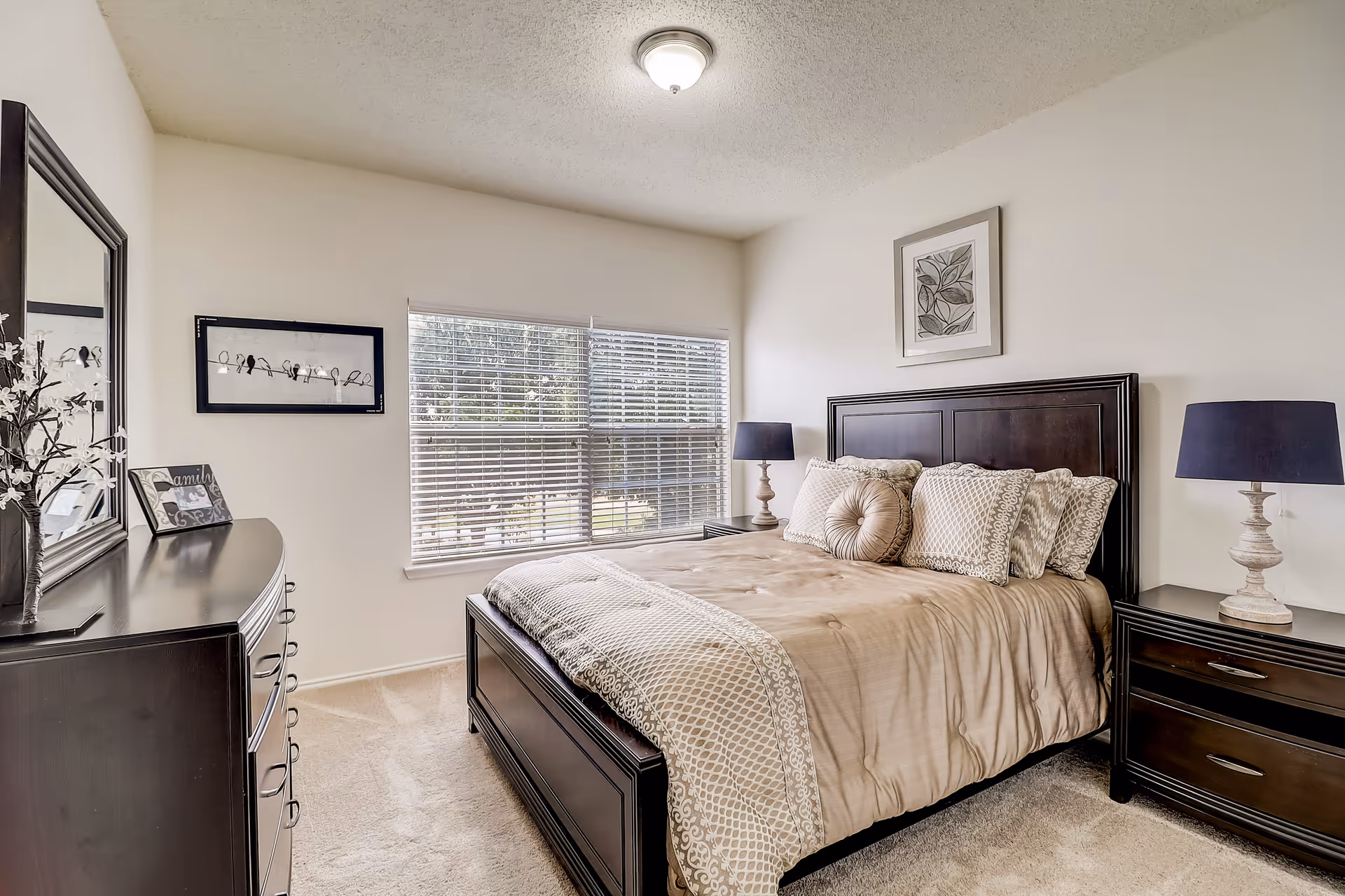 A neatly made bed with beige and patterned bedding in a bedroom. The room has a large window with blinds, two dark wooden nightstands with matching lamps, a dark wooden dresser with a mirror, and framed artwork on the walls. The carpet is light-colored and the walls are painted white.