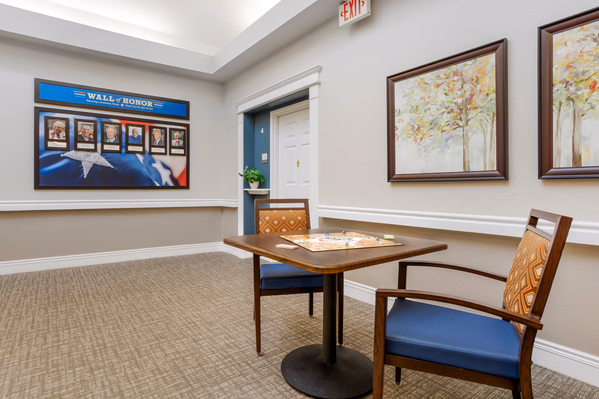 A quiet room in a senior living facility featuring a small square table with two chairs. A board game is set up on the table. On the wall to the left, there is a 'Wall of Honor' display with framed photos of veterans. Two framed paintings of trees hang on the right wall. The room has beige walls, carpeted floor, and a white door with a plant on a small shelf beside it. An exit sign is visible above the door.