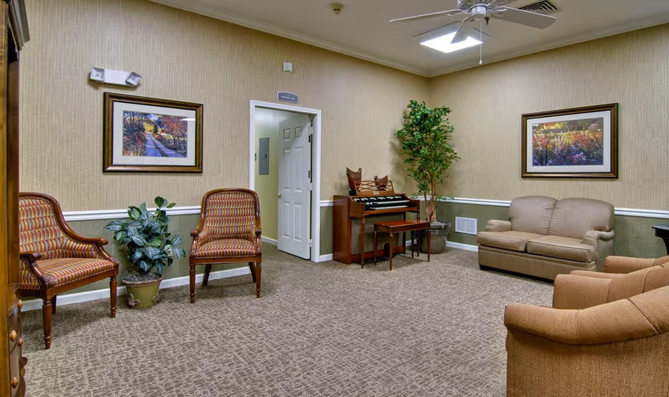 A cozy living room with beige walls and carpet, featuring a ceiling fan, two framed landscape paintings, two patterned armchairs, a beige loveseat, a brown armchair, a potted plant, and an organ with a matching bench near an open door.