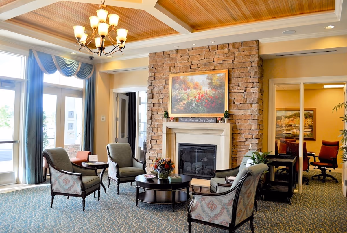 Bright lounge in a senior living facility featuring a stone fireplace, arranged armchairs around a coffee table, chandelier, and large windows.