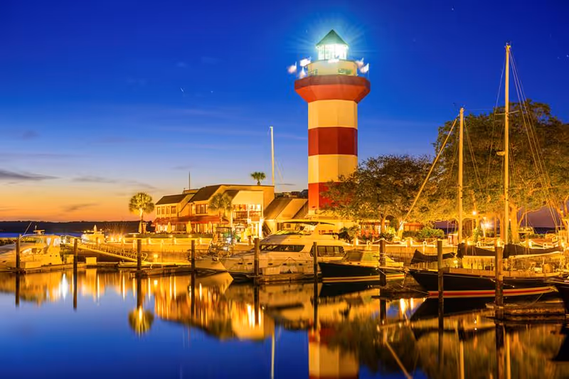 A red-and-white striped lighthouse beside a lit marina with boats reflected on calm water at dusk.